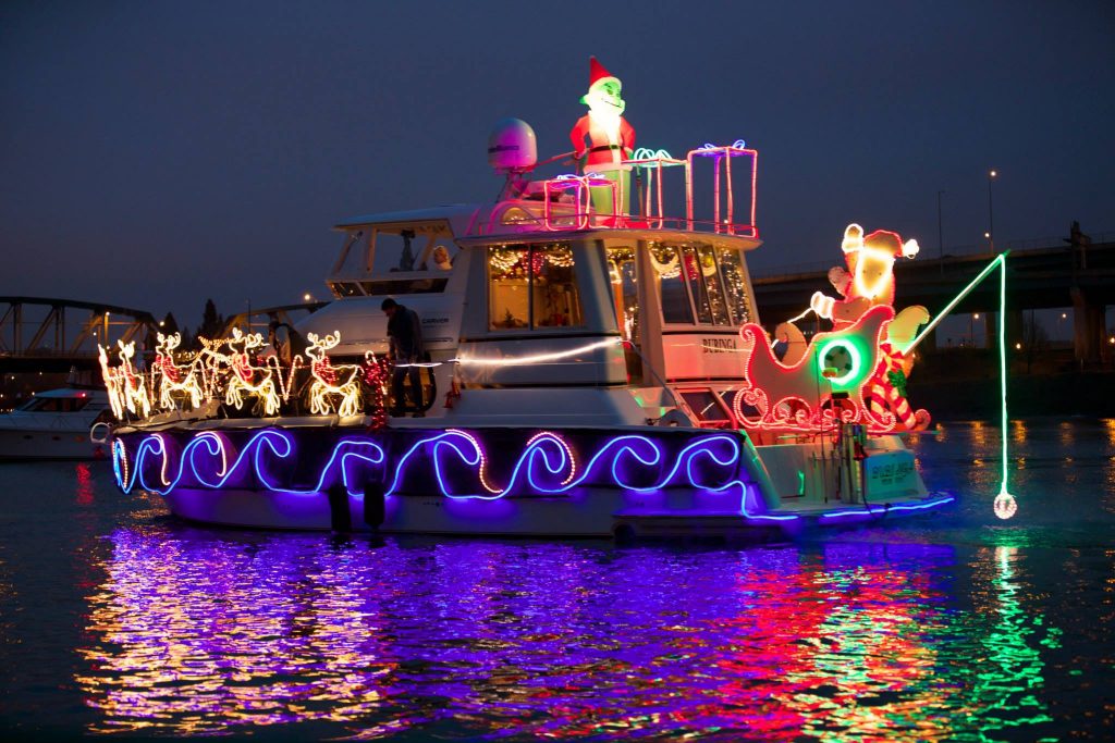 A boat decorated with Christmas lights and blow up holiday decorations in the water at night time. The lights reflect off of the surface of the water.