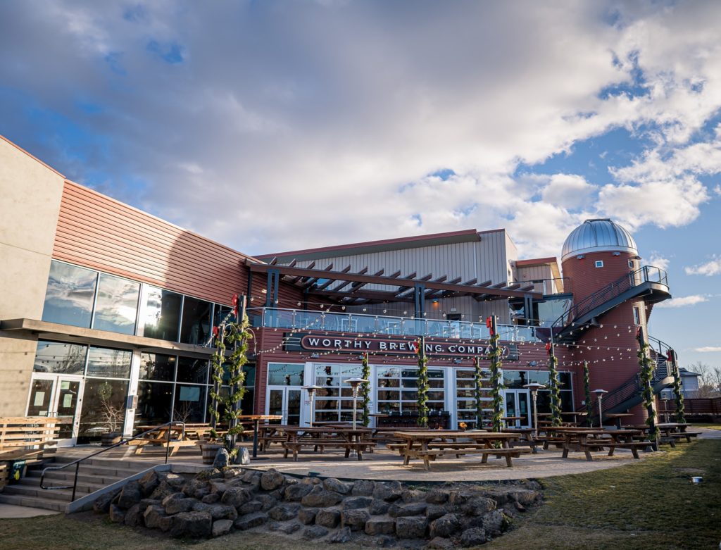 The outside of Worthy Brewing Co in Bend, Oregon.  There's a large outdoor seating area with picnic tables. T he building is tall and modern looking with steel beams, concrete walls and red siding.  Part of the roof looks like an observatory dome made of metal.  There are string lights over the outdoor seating area.  It looks cozy and fun.