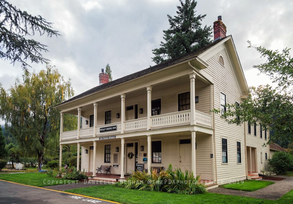 The outside of the Wolf Creek Inn on a cloudy day. It's a two story white building surrounded by green lawn, several plants, and a few trees. There's a large covered front porch, and covered second porch stretching across the inn held up by round white pillars.