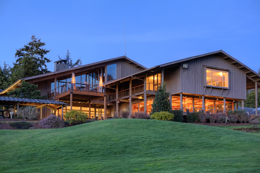 The outside of Salishan Coastal Lodge at night, with warm lights spilling out the windows. The building has wooden siding and looks very nice.