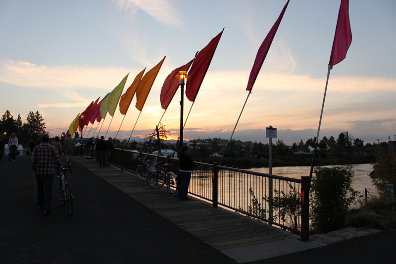 A footbridge goes over the river in Bend at sunset.  People are walking across it.