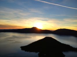 Crater Lake at sunrise. Wizard Island and the rim of the lake are cast in a sharp black silhouette as the sun peeks up over the rim.