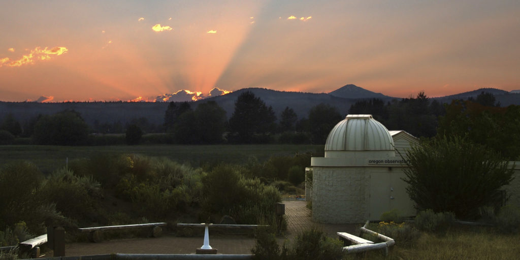 The observatory in Sunriver at sunset.  Rays of sun peak out over low mountains in the distance.  There are trees, a field, and desert brush around the small white observatory building.  There are also wooden benches in a circle outside the building.