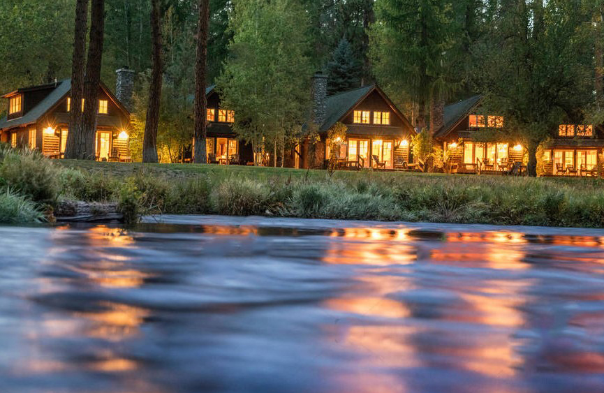 Cabins in the evening on the Metolius River. There's warm light spilling out of the cabins, best oregon towns, spring road trip, best towns to visit, 2024