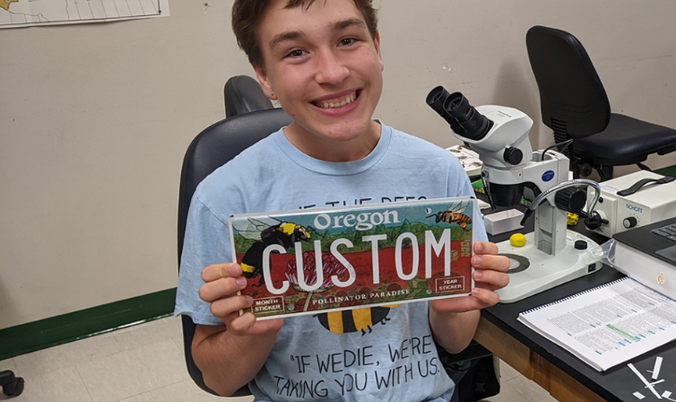 The artist who designed the pollinator license plates holds the plate up with a smile. It's a high school artist.