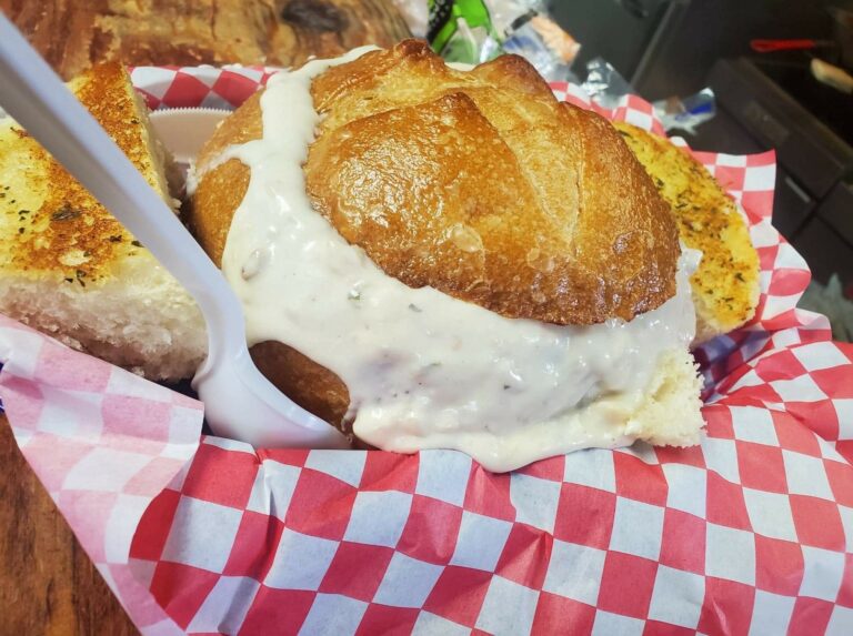 A crispy bread bowl full of thick clam chowder at The Hungry Clam. This dish looks really good. It's in a basket with red and white checkered paper.
