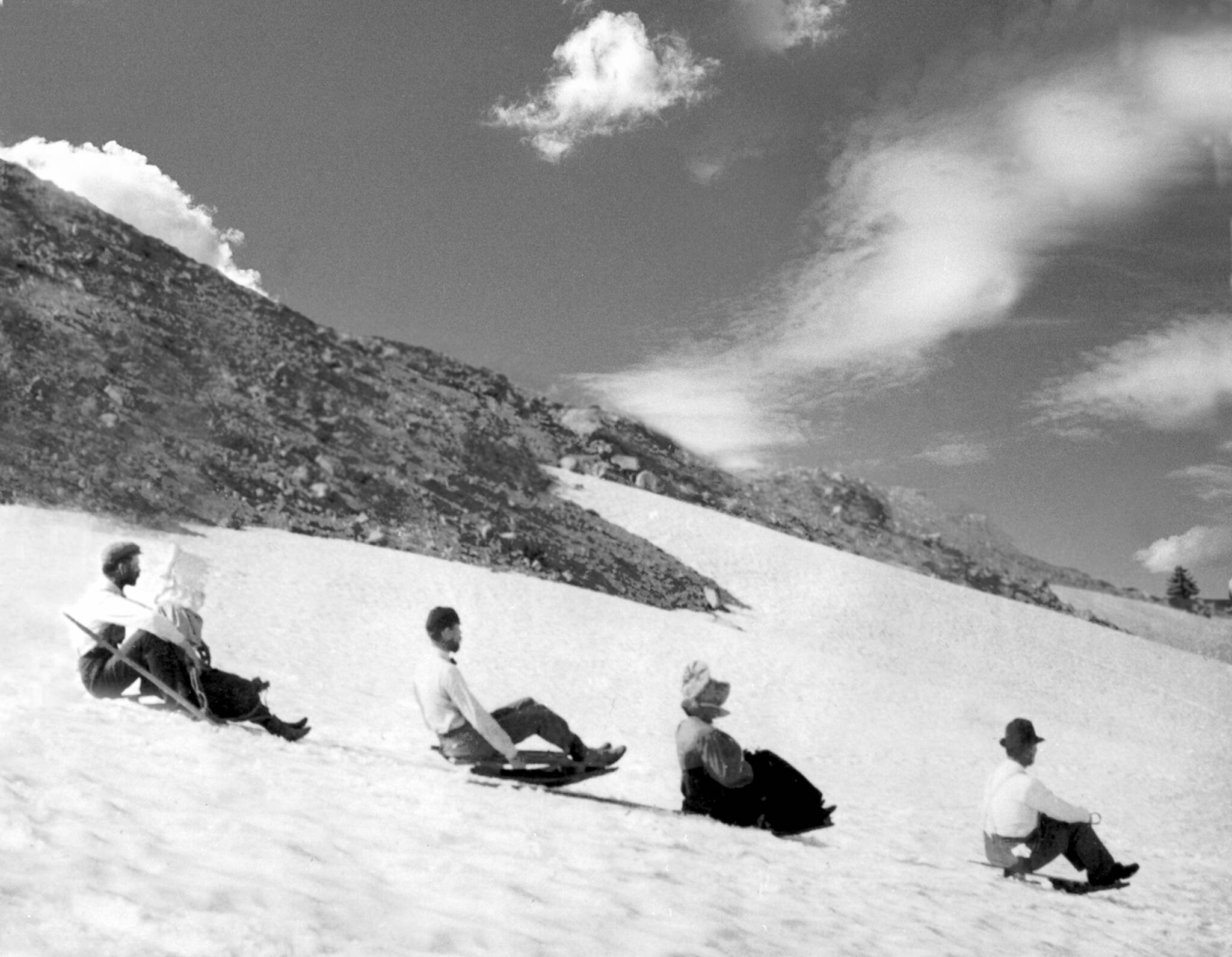 A black and white historical photo of showshoers sitting on the snow in a line at Cooper Spur.