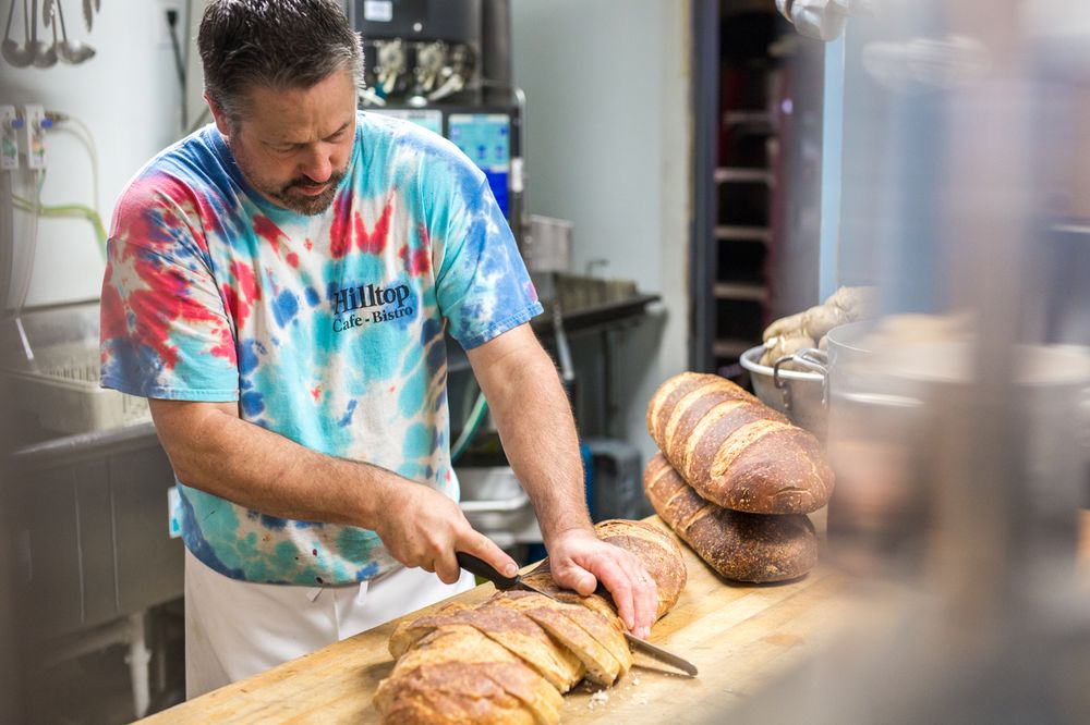 A chef in a blue and red tie dye shirt cuts up fresh loaves of bread.