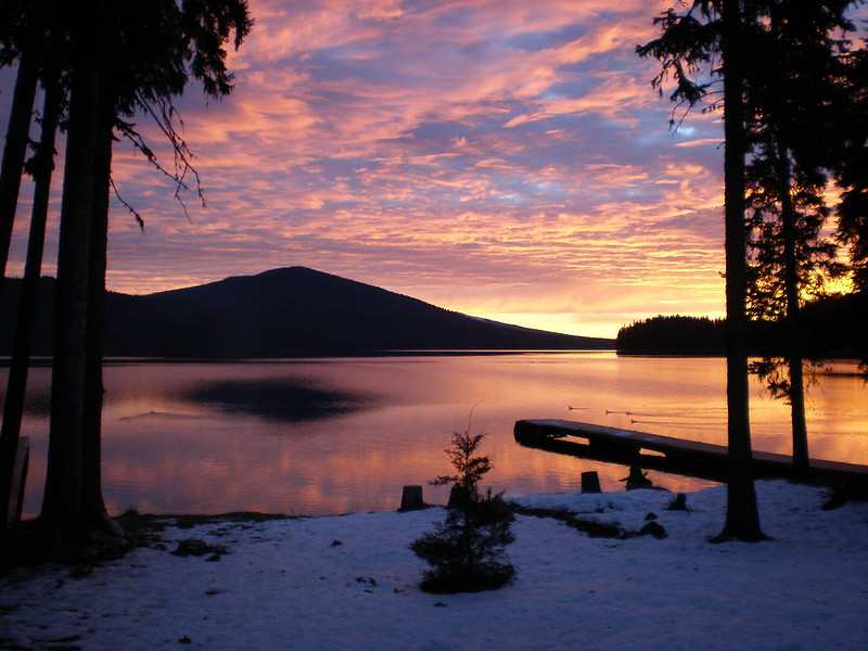 Odell Lake at dawn. The sunset is casting bright pink and orange into the still waters with a small mountain at the edge of the lake.