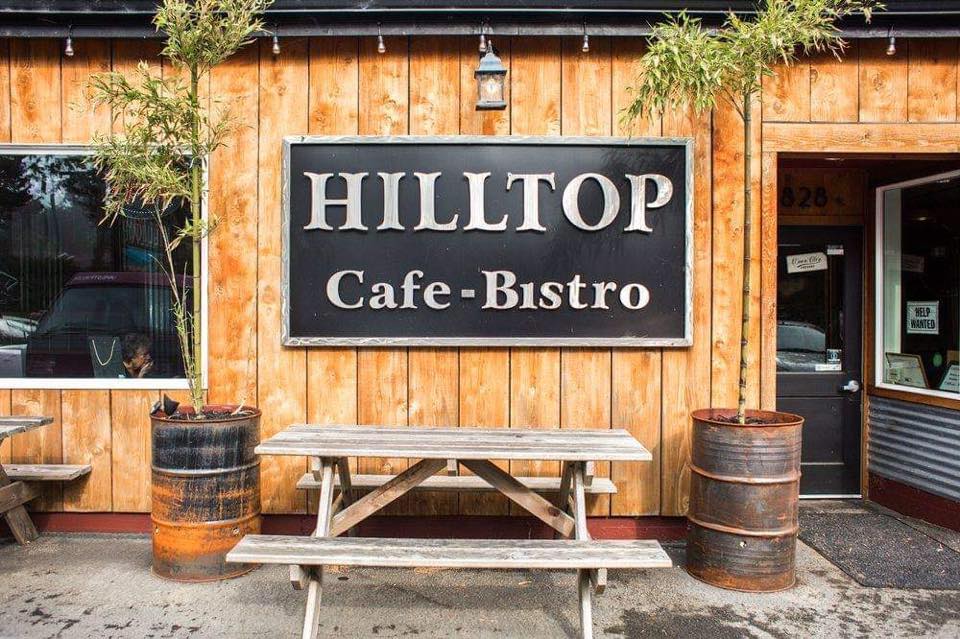The outside of Hilltop Cafe Bistro.  A warm wood wall with a picnic table and two trees in metal barrels.