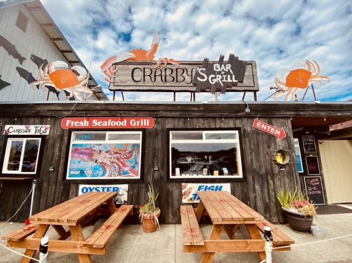 The outside of Crabby's Bar And GRill. It has wood siding, nice wooden signs, accents of red, blue and orange. There are painted crabs and two wooden picnic tables.