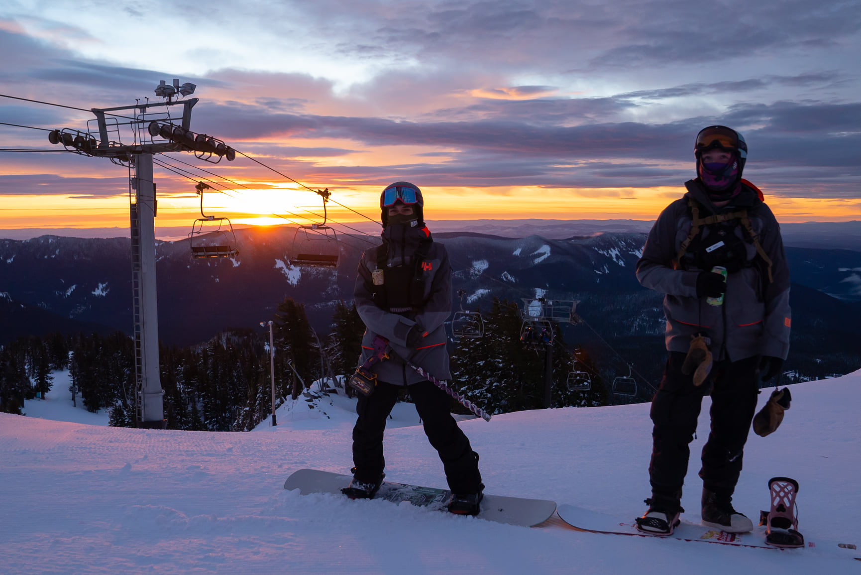 Two snowboarders pose in front of the chairlift with an orange sunset behind them at Mount Hood Meadows.