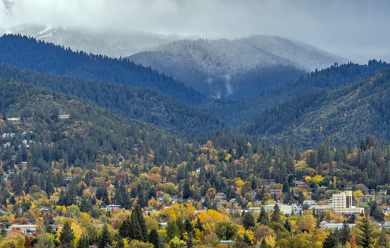An aerial view of Ashland in the fall. There are thousands of vibrant orange and yellow trees, mountains in the background, and a dusting of snow in the mountains. It's very pretty.