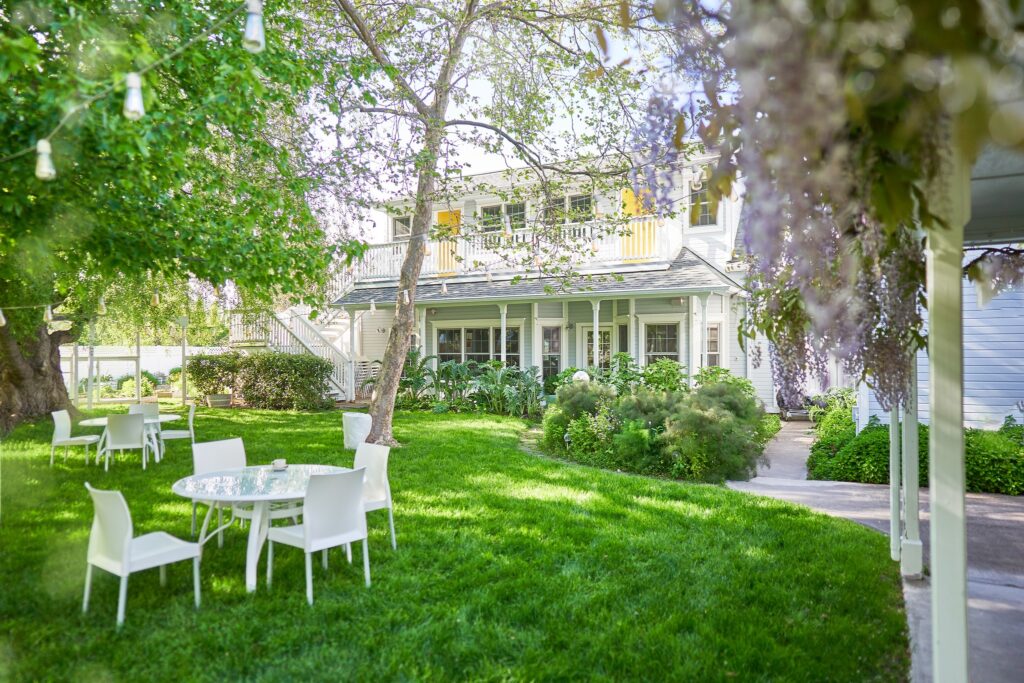 A shaded seating area with a white table and chairs on a green lawn with a white house in the background.