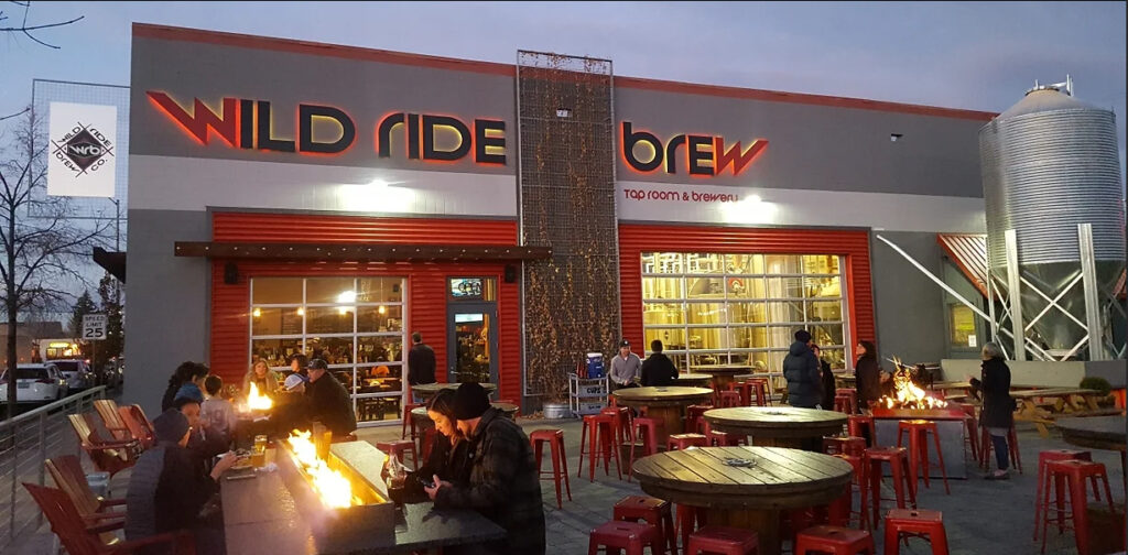 The outside of Wild Ride Taproom. A hip gray building with lots of red accents and outdoor seating in the evening.