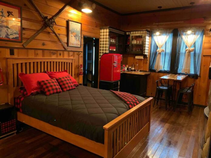 A wood bed with a dark gray comforter and red and black checkered accents. There are wood walls. It's very cozy looking.