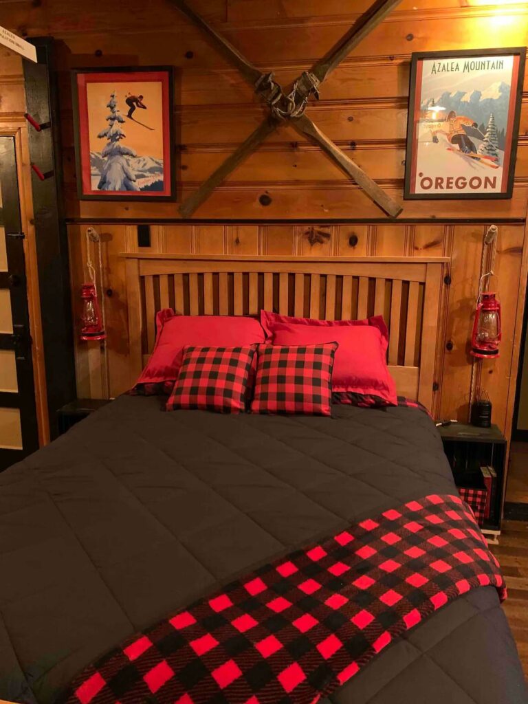 A wood bed with a dark gray comforter and red and black checkered accents. There are wood walls. It's very cozy looking.