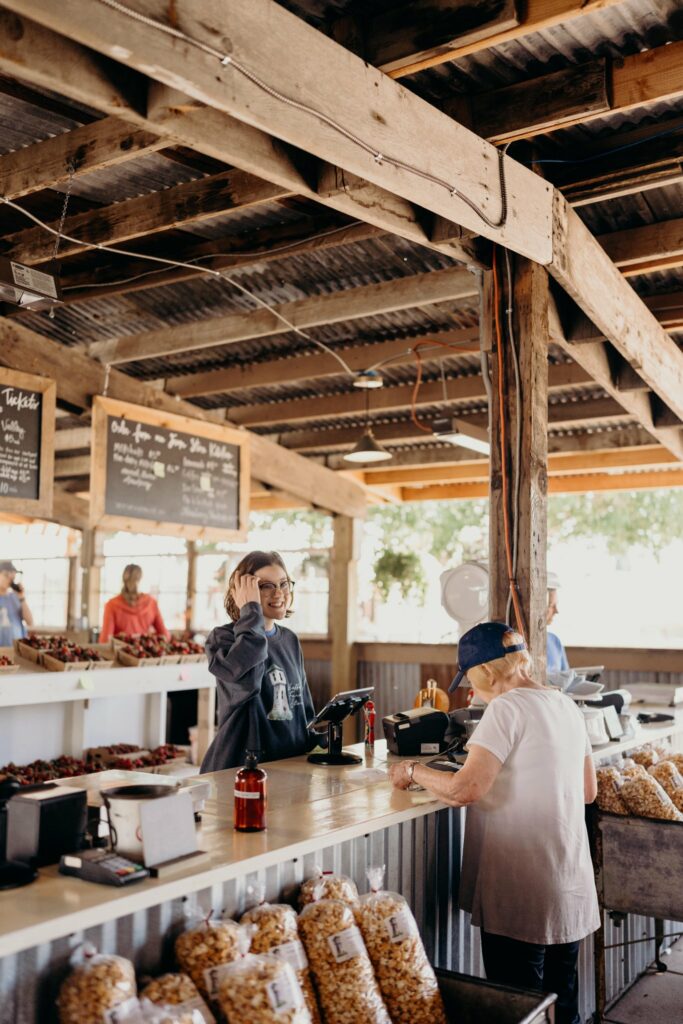 A smiling woman serves a boy with blond hair and a blue hat in an outdoor style farm market at Hoffman Farms. There are berries on the counter, and the market is covered by a structure with wood beams, providing shade.