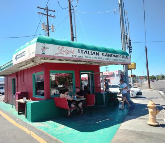 The outside of Luigi's. It's a small red building with a green roof and green painted small outdoor seating area.