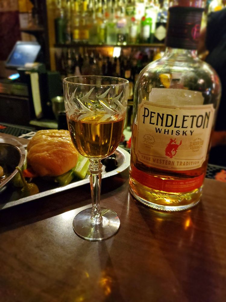 A bottle of Pendleton Whiskey on a wood table. There's also a glass of whiskey and a plate of food.