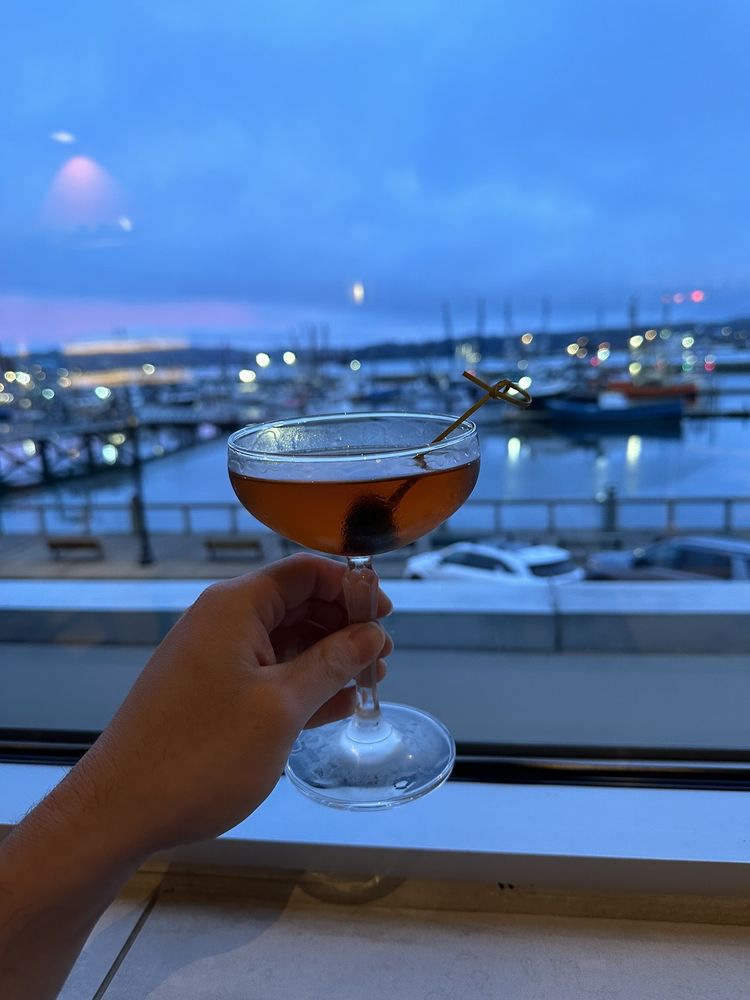 Someone holding up an alcoholic drink in front of the window overlooking the bay and docks in the evening. You can see ships docked in the background.