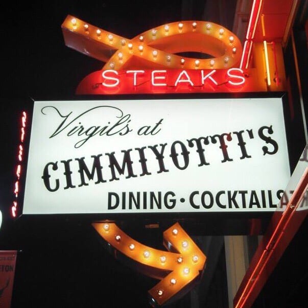 The Virgil's At Cimmiyotti's sign lit up bright at night. It has a glowing orange arrow and red neon words proclaiming: Steaks!