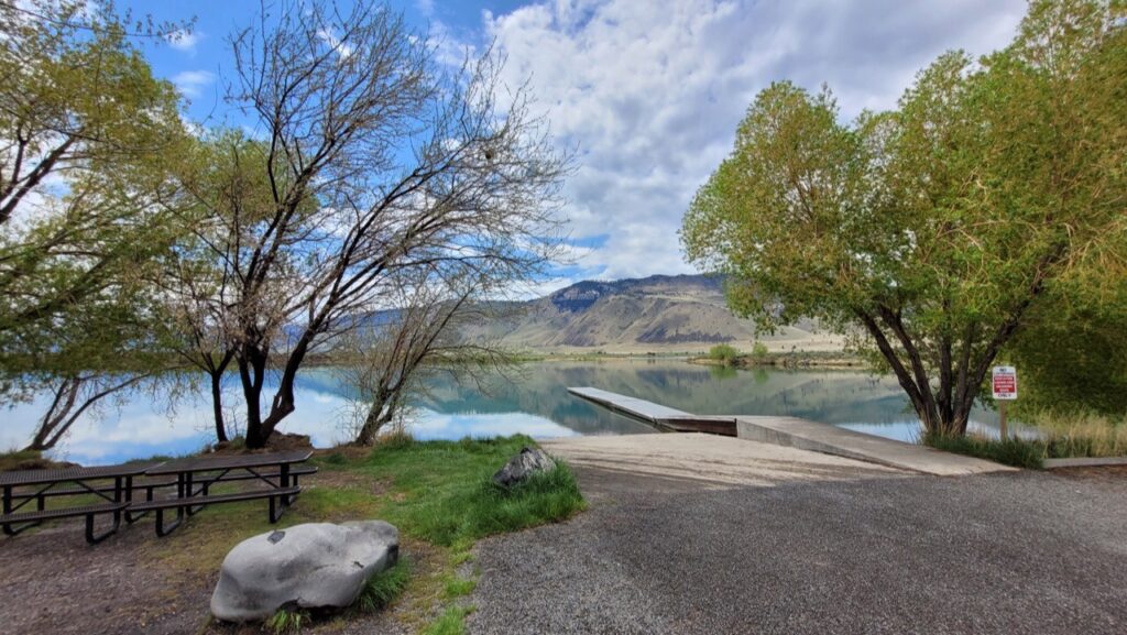 A dock goes down into Ana Reservoir on a nice day.  There are a few trees, some grass, and a picnic bench on the shore of the still waters of the reservoir.