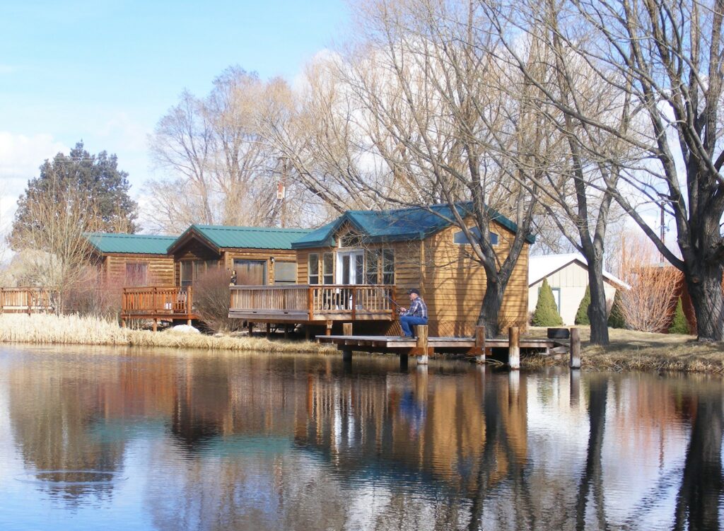 Cute little cabins with green roofs sit right on the edge of a body of calm water. Someone is fishing from the porch of a cabin.