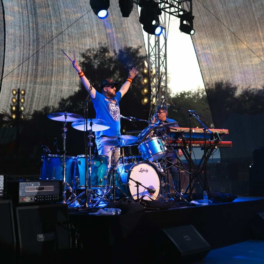The drummer throwing his arms up in the air behind his drums on stage.