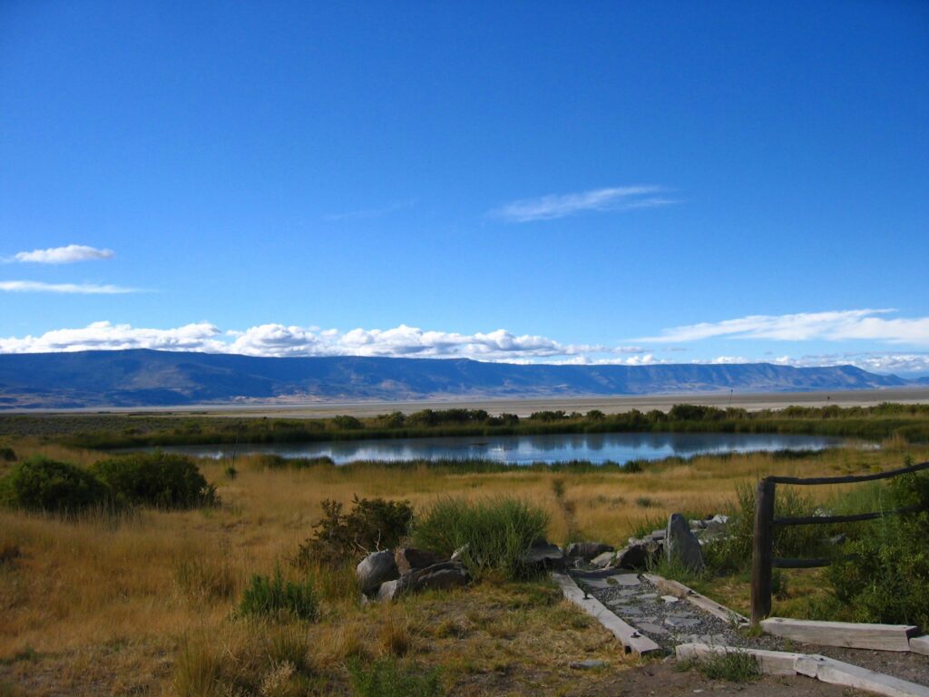 Blue skies reflect in a pool of water. There is a rock path down to the pool of water and low flat desert mountains in the background.