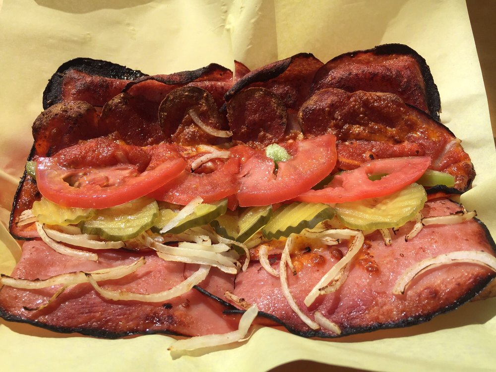 A big sandwich with onions, pickles, tomatoes, and several types of toasted deli meats.