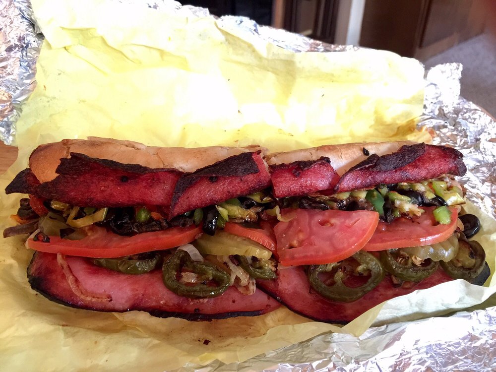 A big sandwich with lots of types of deli meat, pickles, olives, and tomatoes.