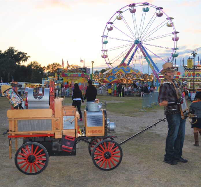 A man pulling a wagon stands in front of the Ferris wheel at the county fair.