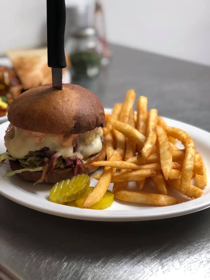 Corned beef brew ben burger with fries, pickles on the plate and a steak knife through the top of the burger.