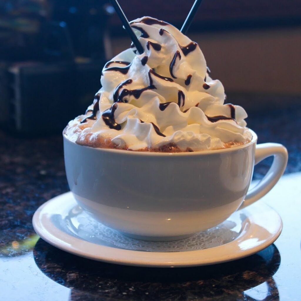 Hot coco with whip cream and a chocolate drizzle in a white mug on a white saucer.
