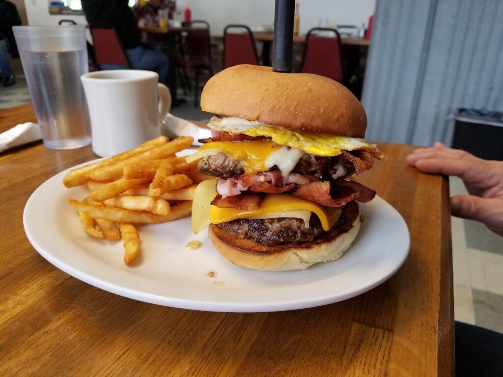 A huge burger with an egg on top, fries on the plate, and a steak knife through the top of the burger.