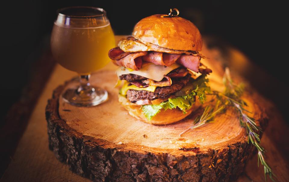 The Big Daddy burger piled high with meat and toppings sitting on a log round next to a glass of alcohol.