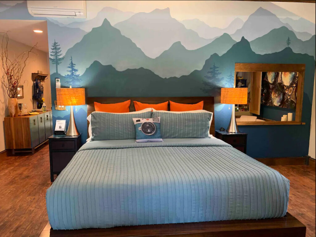 A king size bed with a light blue comforter and orange pillows. The wall behind the headboard is painted like the silhouettes of mountains in shades of blue.