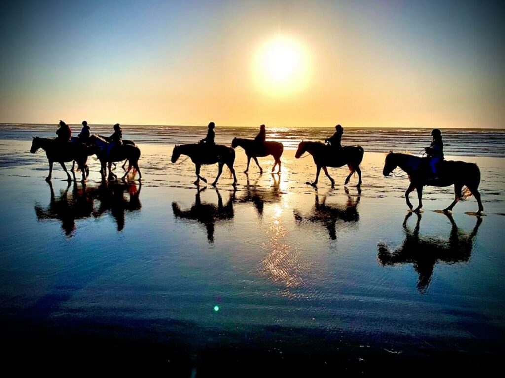 A group of people ride horses along the beach silhouetted by the sunset in Bandon Oregon.