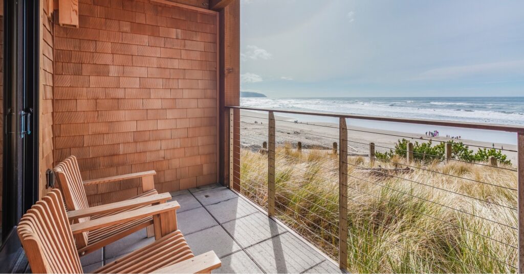 A porch with two wooden chairs overlooking the ocean. Sand and coastal grass are just below the porch.