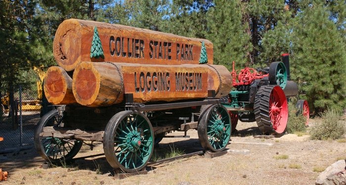 Large logs carved with the words 'Collier State Park Logging Museum' sit on an old train bed.