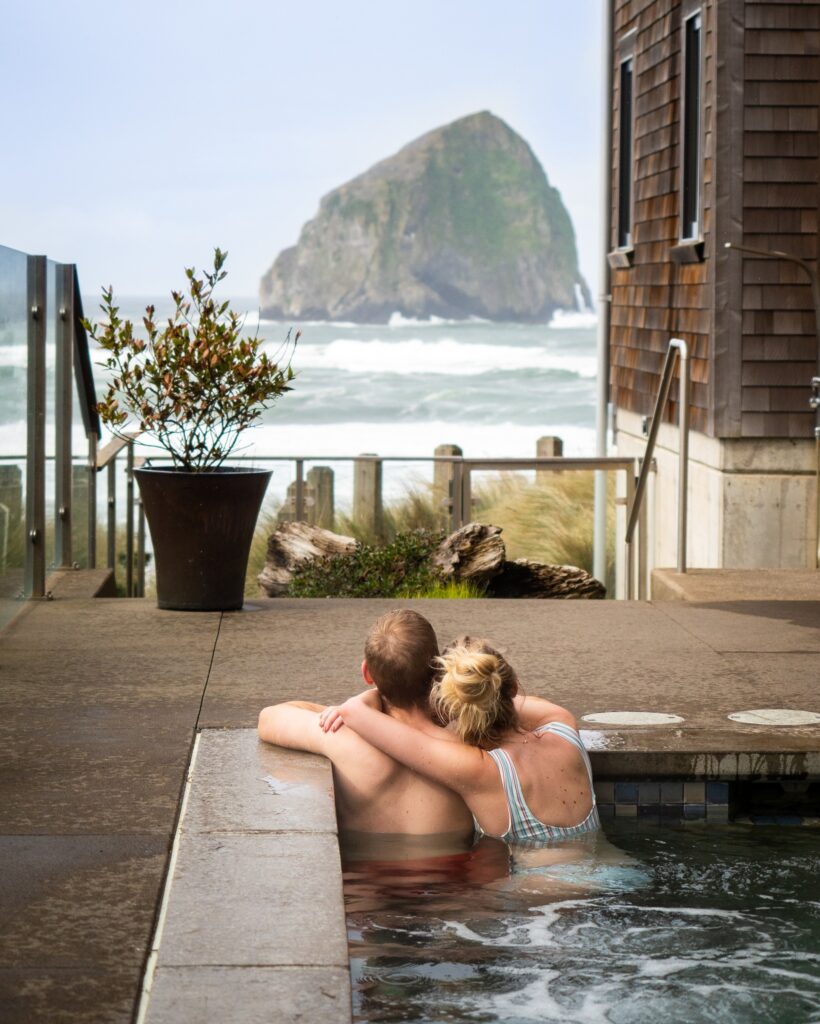 Two people hug in the water at the spa while overlooking Haystack Rock and the ocean views.