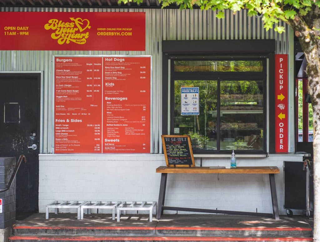 The walkup order window at Bless Your Heart Burgers. The building has metal siding and bright red signs with yellow lettering.