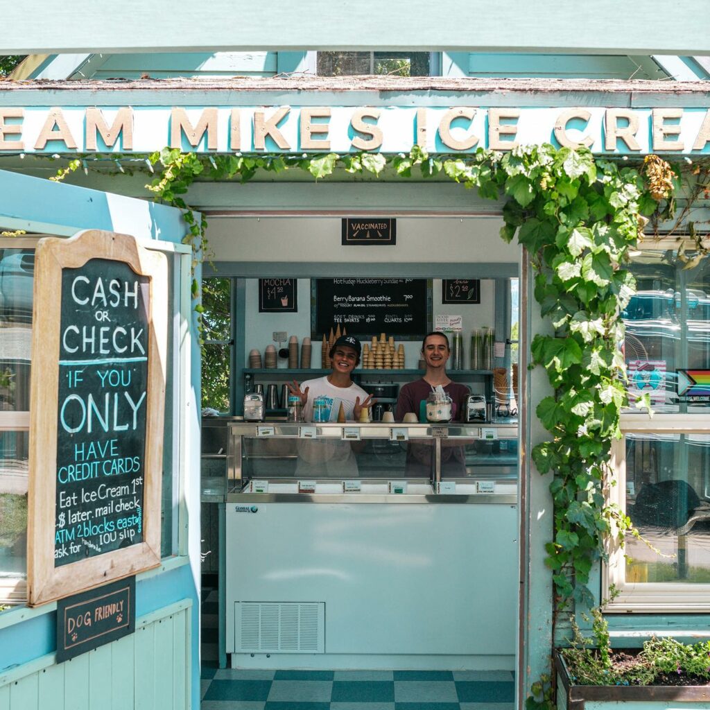 The outside of the cute Mike's Ice Cream shop. The building looks like a house and is a cute sage or aqua green color. Two people smile and wait to serve customers at a walk up window.
