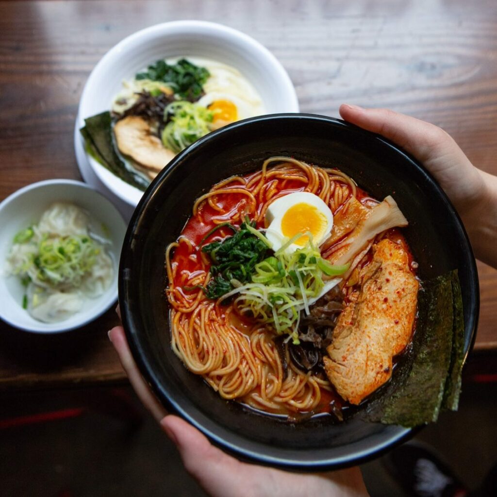 A pair of hands holds a black bowl of red ramen with vegetables and a boiled egg. Two white bowls of food sit on a table behind it.