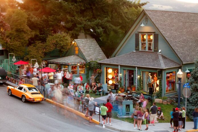 A zoomed out view of Mike's Ice Cream and the business next door. Both look like green houses, and there is a crowd of people outside on the lawn and lounging in chairs eating ice cream.