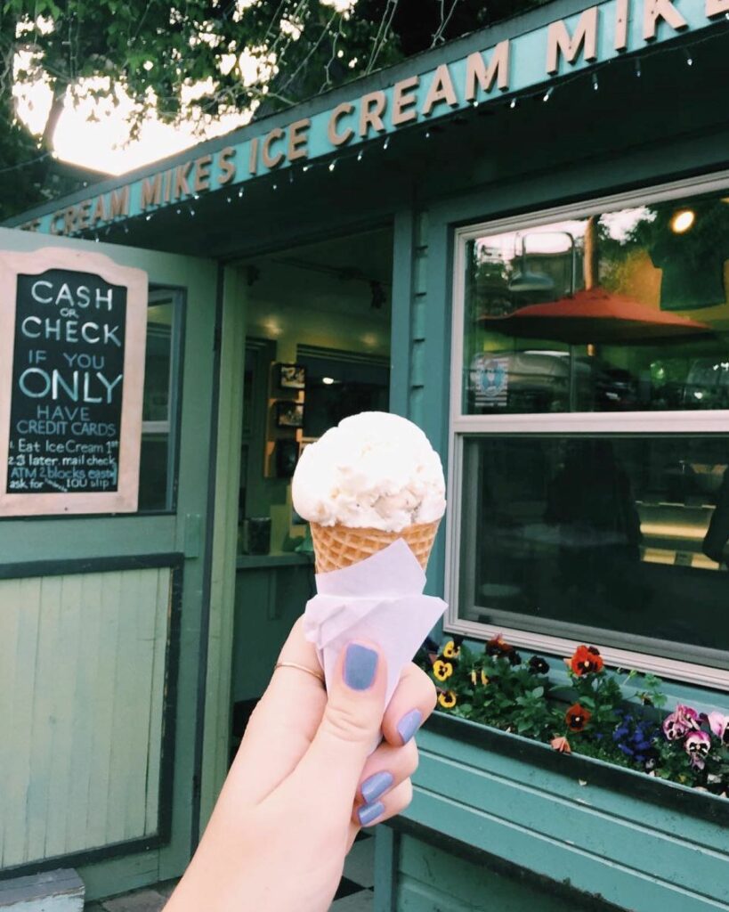 A person holds up an ice cream cone in front of the aqua colored Mike's Ice Cream building.