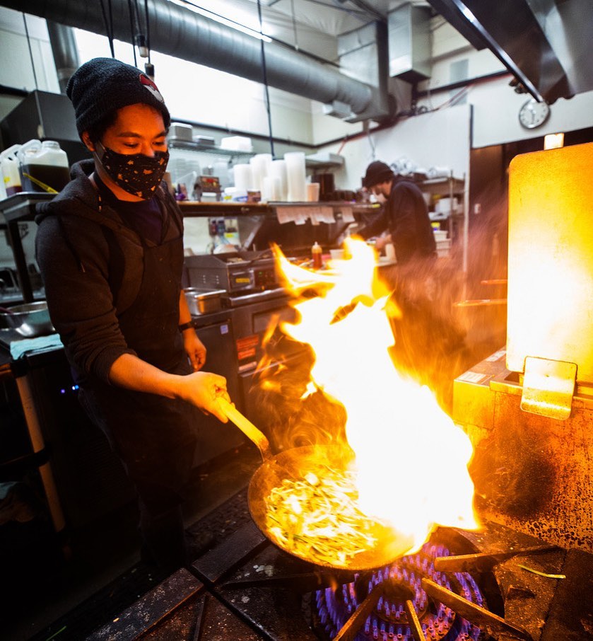 A chef works his magic in the Kinboshi Ramen kitchen. He's cooking something in a pan that has flames.