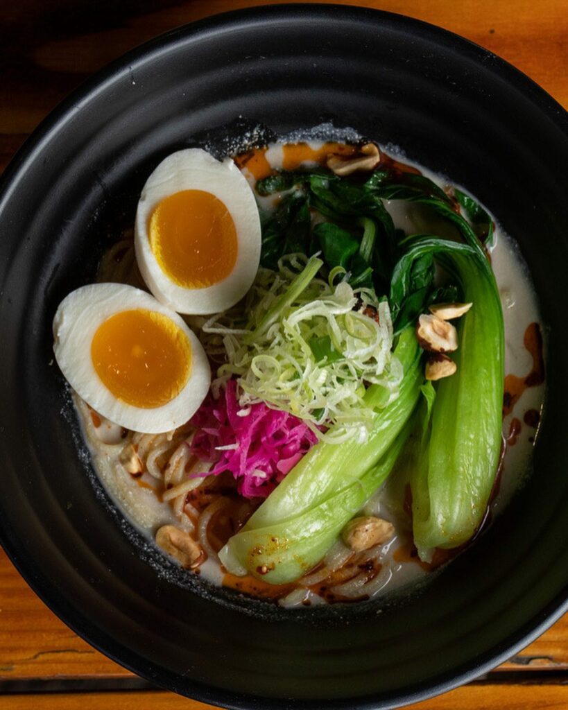 A bowl of ramen packed with green, and purple vegetable toppings and a boiled egg. It looks so good!