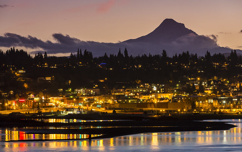 A night time view of Hood River, Oregon as seen from across the Columbia River.  Lights reflect off of the river and Mount Hood looms in the background silhouetted against the late evening sky.